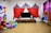 A children's play area with colorful decorations. The room features a large TV with speakers on a wooden stand. Above it is a circus-themed backdrop with red, blue, and yellow fabric resembling tents. To the left, there's a large cluster of balloons in various colors and a life-sized plush Mickey Mouse figure. The floor is strewn with more balloons and cushions. On the right, a rack displays costumes, and there's a small puppet theater booth.