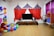 A children's play area with colorful decorations. The room features a large TV with speakers on a wooden stand. Above it is a circus-themed backdrop with red, blue, and yellow fabric resembling tents. To the left, there's a large cluster of balloons in various colors and a life-sized plush Mickey Mouse figure. The floor is strewn with more balloons and cushions. On the right, a rack displays costumes, and there's a small puppet theater booth.