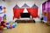 A children's play area with colorful decorations. The room features a large TV with speakers on a wooden stand. Above it is a circus-themed backdrop with red, blue, and yellow fabric resembling tents. To the left, there's a large cluster of balloons in various colors and a life-sized plush Mickey Mouse figure. The floor is strewn with more balloons and cushions. On the right, a rack displays costumes, and there's a small puppet theater booth.
