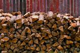 A large stack of cut firewood neatly arranged against a rough, weathered wooden fence. The wood pieces are various sizes, with bark still attached, and display a range of natural browns.