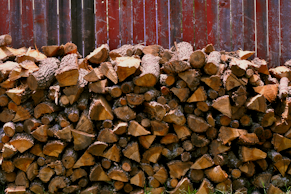 A large stack of cut firewood neatly arranged against a rough, weathered wooden fence. The wood pieces are various sizes, with bark still attached, and display a range of natural browns.