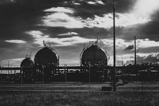 Three large spherical tanks set within an industrial complex, surrounded by barbed wire fencing. The scene is under a dramatic cloudy sky, suggesting late afternoon or early evening lighting. Power lines and utility poles are visible to the right.