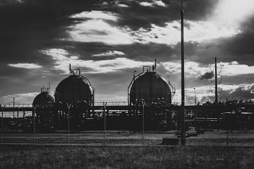 Three large spherical tanks set within an industrial complex, surrounded by barbed wire fencing. The scene is under a dramatic cloudy sky, suggesting late afternoon or early evening lighting. Power lines and utility poles are visible to the right.