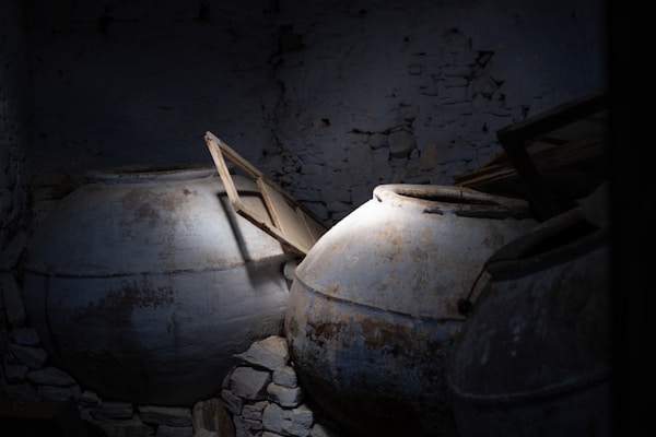 Large, ancient clay pots, partially lit, are surrounded by a dark, textured stone wall. Some old wooden planks are leaning against the pots.