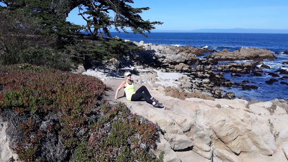 woman in yellow tank top and black pants sitting on rock near body of water during