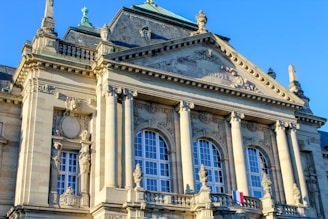 An ornate historical building featuring classical architecture with intricate stone carvings and large arched windows. The facade includes columns and detailed sculptures, as well as a small French flag displayed prominently.