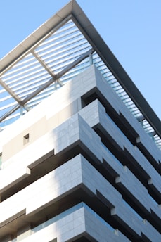 A modern multi-story building featuring a series of protruding balconies and a unique roof structure with metal beams and a glass-covered section. The architecture is sleek and angular, utilizing large blocks of concrete or stone for the exterior. The building is set against a clear blue sky, with light and shadows playing on the surfaces.
