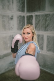 A person stands in a fighting stance wearing boxing gloves, with one glove extended towards the camera. The background features a concrete wall. The individual appears confident and focused, with light-colored hair and wearing a sleeveless athletic top.