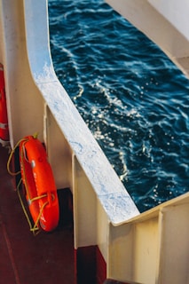 A lifebuoy is affixed to the railing of a boat, visible against the backdrop of the deep blue sea. The sunlight reflects off the waves, creating a sparkling effect on the water's surface.