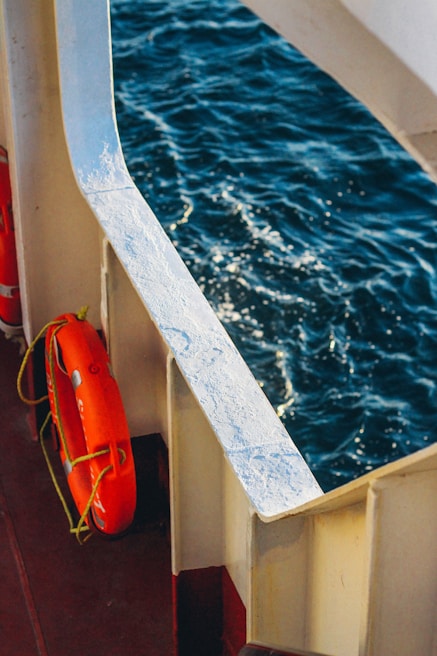 A lifebuoy is affixed to the railing of a boat, visible against the backdrop of the deep blue sea. The sunlight reflects off the waves, creating a sparkling effect on the water's surface.