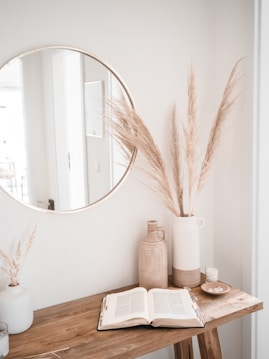 A minimalist interior scene featuring a wooden table with an open book, a tall vase holding dried pampas grass, and a ceramic jug. A round mirror is mounted on the wall, reflecting part of the room and enhancing the sense of space. The setting is calm and natural, with neutral tones and simple decorative elements.