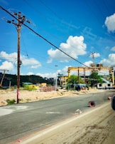 A construction site with large cranes and industrial machinery is visible. The site is bordered by a paved road, with some debris and construction materials scattered around. Overhead, several power lines stretch across a clear blue sky with scattered fluffy white clouds.