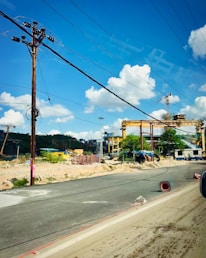 A construction site with large cranes and industrial machinery is visible. The site is bordered by a paved road, with some debris and construction materials scattered around. Overhead, several power lines stretch across a clear blue sky with scattered fluffy white clouds.