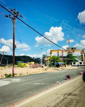 A construction site with large cranes and industrial machinery is visible. The site is bordered by a paved road, with some debris and construction materials scattered around. Overhead, several power lines stretch across a clear blue sky with scattered fluffy white clouds.