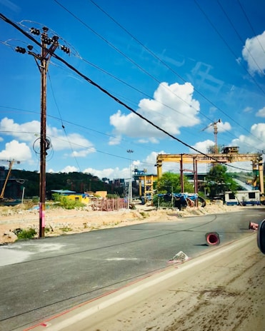 A construction site with large cranes and industrial machinery is visible. The site is bordered by a paved road, with some debris and construction materials scattered around. Overhead, several power lines stretch across a clear blue sky with scattered fluffy white clouds.