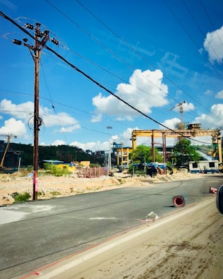 A construction site with large cranes and industrial machinery is visible. The site is bordered by a paved road, with some debris and construction materials scattered around. Overhead, several power lines stretch across a clear blue sky with scattered fluffy white clouds.