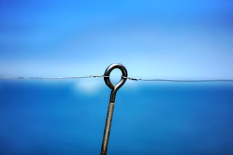 A metal hook is mounted in front of a blurred blue background, appearing to be hanging vertically with a thin wire looped through it.