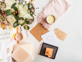 A flat lay featuring a bouquet of white roses and greenery, placed on a white surface alongside a lit candle, a light pink fringed cloth, and several tags with circular text designs. Additional objects include a geometric candle, a pile of books with visible titles and a wooden stamp resting on an ink pad.
