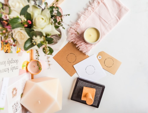 A flat lay featuring a bouquet of white roses and greenery, placed on a white surface alongside a lit candle, a light pink fringed cloth, and several tags with circular text designs. Additional objects include a geometric candle, a pile of books with visible titles and a wooden stamp resting on an ink pad.