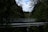 A metal bridge and a pipeline cross over a forested area with dense evergreen trees, under a partly cloudy sky.