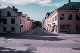 A quiet, empty street lined with two-story buildings featuring a European architectural style. The buildings are painted in soft pastels with several windows and small storefronts. One prominent sign reads 'The English Bookshop'. The sky is partly cloudy, and shadows are cast on the street from the trees lining the sidewalks.