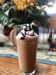 A glass filled with a creamy chocolate beverage topped with whipped cream and a drizzle of chocolate syrup. It is placed on a wooden table with a blurred background featuring plants and colorful decor.