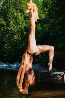 A person is performing aerial silk acrobatics outdoors, suspended upside down in a natural setting. The acrobat's body is gracefully extended, with one leg pointed upwards and hands holding onto orange silks. Surrounded by lush greenery and a shallow body of water, the scene evokes a sense of serenity and strength.