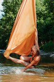 A person is engaged in aerial silk performance over a body of water, with vibrant green trees in the background. The performer hangs gracefully from a large, flowing orange fabric, with one leg extended and toes pointed, creating a dynamic and elegant pose. Sunlight filters through the trees, highlighting the colors and motion.