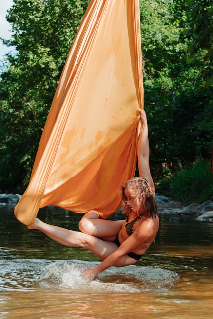 A person is engaged in aerial silk performance over a body of water, with vibrant green trees in the background. The performer hangs gracefully from a large, flowing orange fabric, with one leg extended and toes pointed, creating a dynamic and elegant pose. Sunlight filters through the trees, highlighting the colors and motion.