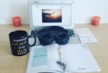 A workspace setup on a wooden desk featuring a laptop displaying a webpage with an image of an airplane wing against a sunset sky. Black headphones rest on the laptop keyboard. Nearby, a black coffee mug with gold and white text sits next to a notebook opened to a checklist and some handwritten notes. Two pink candles are placed symmetrically on either side.