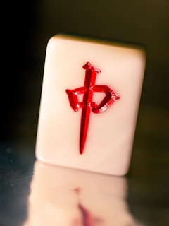 A mahjong tile with a bright red Chinese character or symbol highlighted against a pale, glossy surface. The tile is positioned upright, casting a reflection onto a smooth surface below. The background is dark and slightly out of focus.