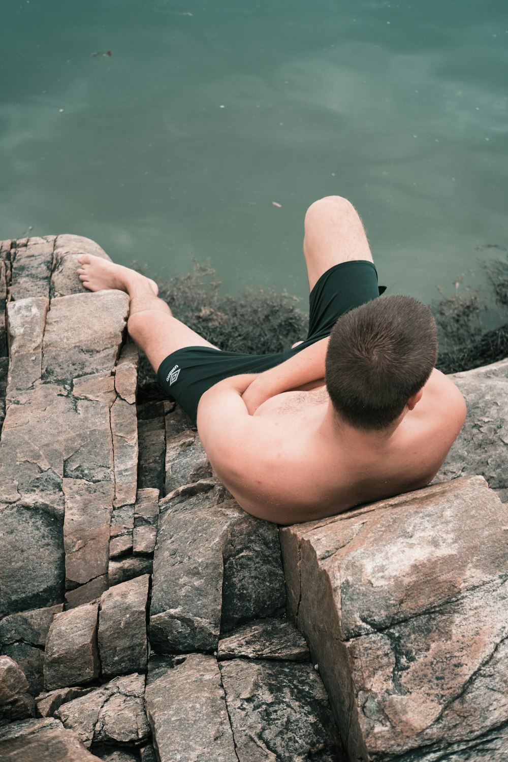 Hombre en pantalones cortos negros sentado en una roca de concreto gris cerca del cuerpo de agua durante el día