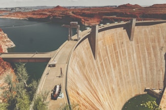 A massive concrete dam spans across a deep valley surrounded by rugged red earth terrain. The reservoir behind the dam contains a large body of dark water, contrasting with the arid, desert-like environment. There is a bridge and some structures on top of the dam, with machinery and a few scattered pieces of equipment visible next to it. Sparse vegetation is seen in the foreground.