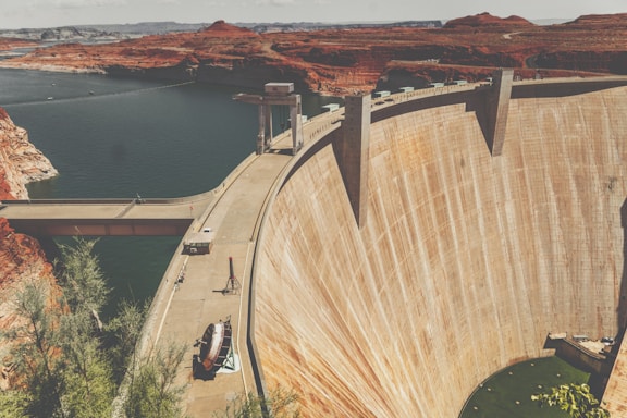 A massive concrete dam spans across a deep valley surrounded by rugged red earth terrain. The reservoir behind the dam contains a large body of dark water, contrasting with the arid, desert-like environment. There is a bridge and some structures on top of the dam, with machinery and a few scattered pieces of equipment visible next to it. Sparse vegetation is seen in the foreground.
