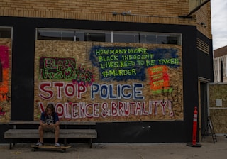 A boarded-up wall features large, vibrant graffiti with powerful social justice messages. The phrases 'Black Lives Matter' and 'Stop Police Violence & Brutality!' are prominently displayed in various bright colors, including red, blue, and green. Nearby, someone is seated on a bench looking at a phone, with a skateboard underneath their feet. A construction cone is placed on the sidewalk beside them.