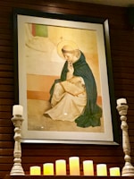 A framed artwork of a religious figure is displayed on a wall. The figure is depicted with a halo, wearing a robe, and appears to be in contemplation or prayer while holding a book. Below the painting, several lit candles and two ornate candleholders enhance the serene and sacred atmosphere.