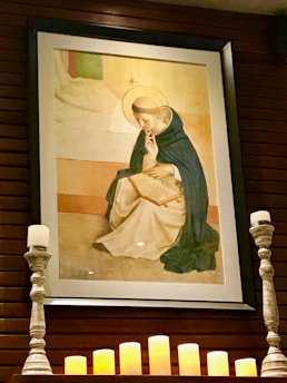 A framed artwork of a religious figure is displayed on a wall. The figure is depicted with a halo, wearing a robe, and appears to be in contemplation or prayer while holding a book. Below the painting, several lit candles and two ornate candleholders enhance the serene and sacred atmosphere.