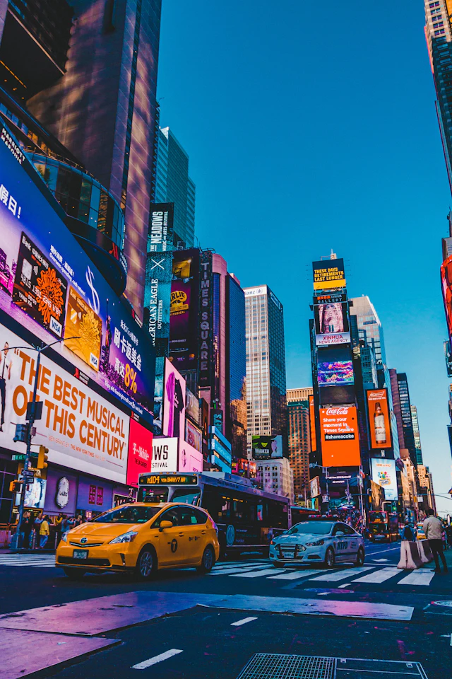 Street-level view of Times Square's neon billboards with a yellow taxi crossing in daylight.