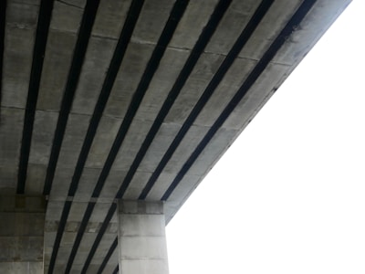 A close-up view of the underside of a concrete bridge featuring longitudinal beams and a vertical support column. The structure appears massive and industrial, with the beam alignment creating a pattern.