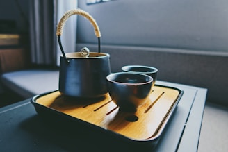 A dark metallic teapot with a woven handle sits on a wooden tray beside two matching cups. The setting appears to be indoors, possibly in a cozy or modern dining area with a soft light illuminating the teaware, creating a calm atmosphere.
