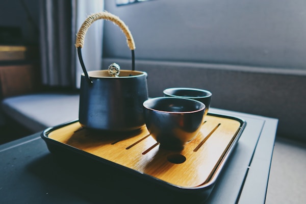 A dark metallic teapot with a woven handle sits on a wooden tray beside two matching cups. The setting appears to be indoors, possibly in a cozy or modern dining area with a soft light illuminating the teaware, creating a calm atmosphere.