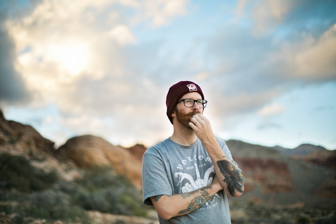 A tattooed man staring into the distance contemplating and thinking with desert hills and clouds behind him.