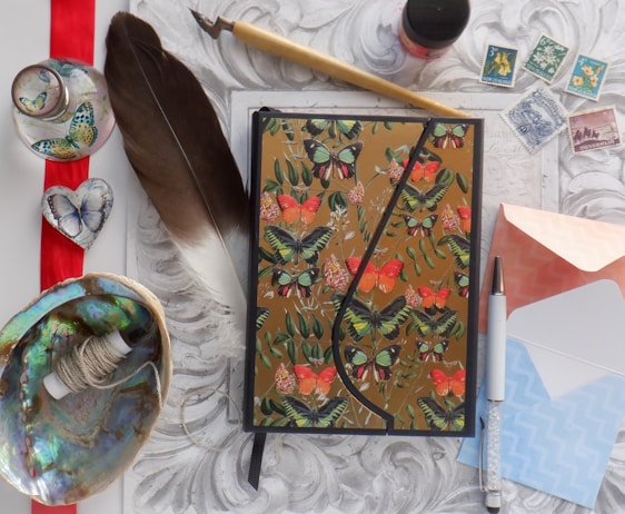 An assortment of artistic items arranged on a decorative surface includes a notebook with butterfly designs, a large brown feather, an ink pen with a sparkly exterior, small envelopes in pastel colors, vintage postage stamps, a bowl with a spool of thread, a heart-shaped butterfly-themed ornament, a glass bottle with butterfly motifs, and a decorative writing quill.
