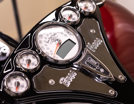 Close-up view of a motorcycle dashboard featuring multiple gauges and meters with chrome accents. The design includes a prominent speedometer with a white face and red needle, accompanied by smaller temperature and fuel gauges. The glossy black finish and metallic details add a sleek and polished look.