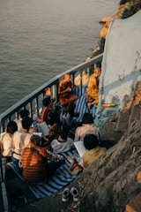 A group of people, including monks, are gathered on a small balcony or terrace by the water. They are sitting on mats and appear to be engaged in study or meditation. The setting sun casts warm, golden light over the scene, highlighting the serene and contemplative atmosphere.