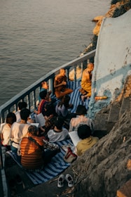A group of people, including monks, are gathered on a small balcony or terrace by the water. They are sitting on mats and appear to be engaged in study or meditation. The setting sun casts warm, golden light over the scene, highlighting the serene and contemplative atmosphere.