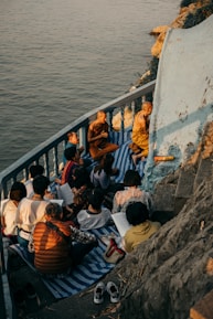A group of people, including monks, are gathered on a small balcony or terrace by the water. They are sitting on mats and appear to be engaged in study or meditation. The setting sun casts warm, golden light over the scene, highlighting the serene and contemplative atmosphere.