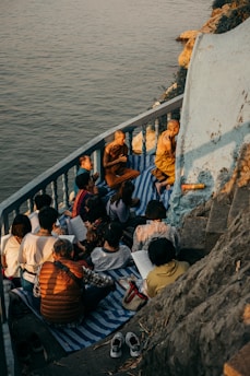 A group of people, including monks, are gathered on a small balcony or terrace by the water. They are sitting on mats and appear to be engaged in study or meditation. The setting sun casts warm, golden light over the scene, highlighting the serene and contemplative atmosphere.