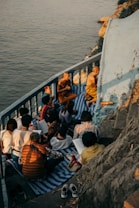 A group of people, including monks, are gathered on a small balcony or terrace by the water. They are sitting on mats and appear to be engaged in study or meditation. The setting sun casts warm, golden light over the scene, highlighting the serene and contemplative atmosphere.