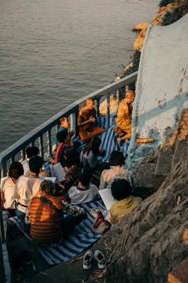 A group of people, including monks, are gathered on a small balcony or terrace by the water. They are sitting on mats and appear to be engaged in study or meditation. The setting sun casts warm, golden light over the scene, highlighting the serene and contemplative atmosphere.
